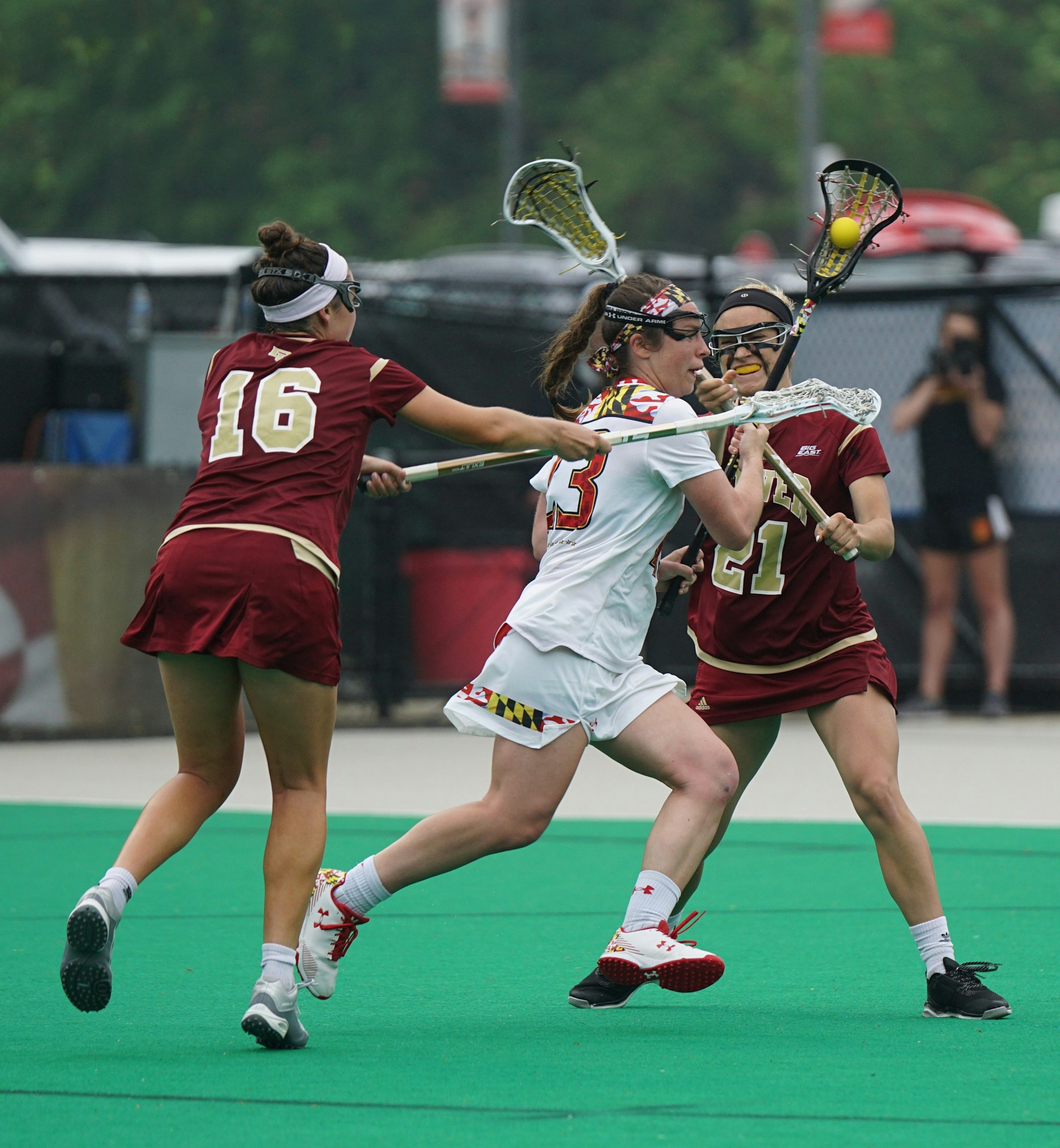 Three women's lacrosse players battling for possession of the ball in the open field.